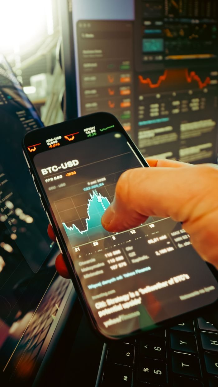 about-01 A hand holds a smartphone displaying BTC-USD chart amidst digital trading screens, highlighting crypto finance.